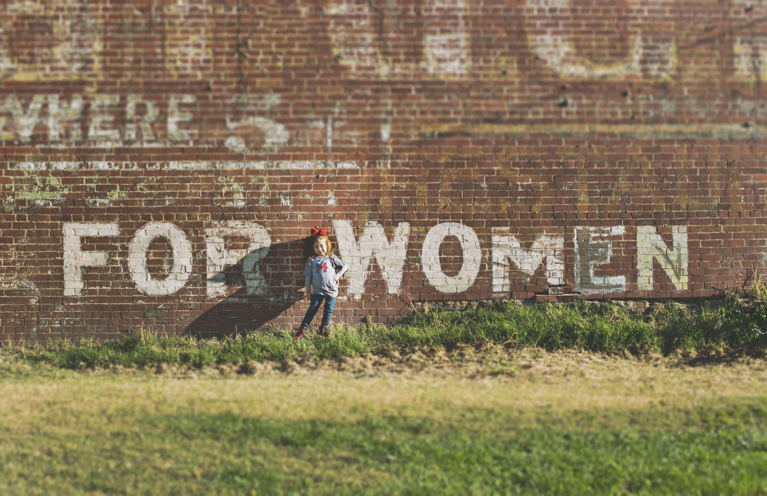 A little girl is standing in front of a brick wall with the words "For Women" painted on it.